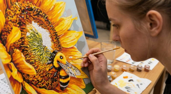 Woman carefully painting a vibrant sunflower and bumblebee artwork on a canvas using a paint by numbers kit, showcasing detailed brushwork, creative expression, relaxing DIY art, colorful floral design, and an enjoyable home crafting experience.